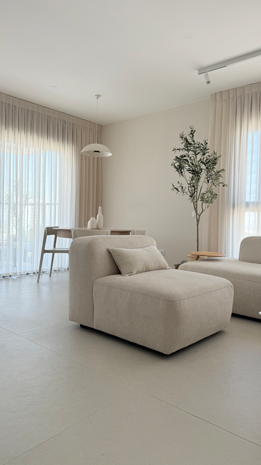 סלון מודרני מעוצב בסגנון מינימליסטי בגווני שמנת עם ספה מודולרית, עציץ זית דקורטיבי ופינת אוכל ברקע.