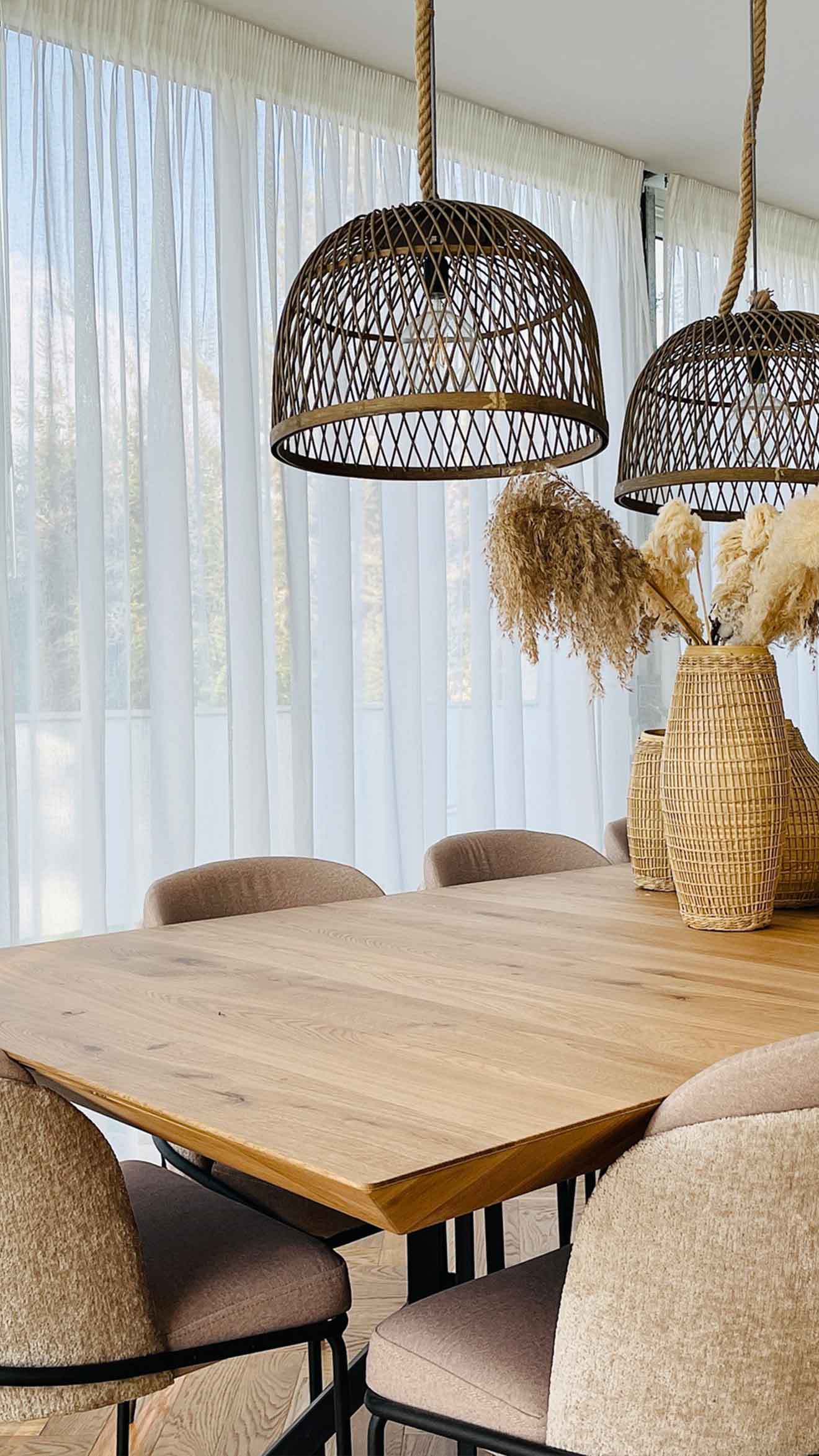 וילון שקוף לבן מעוצב כרקע לפינת אוכל עם שולחן עץ, כורסאות מרופדות וגופי תאורה קלועים.