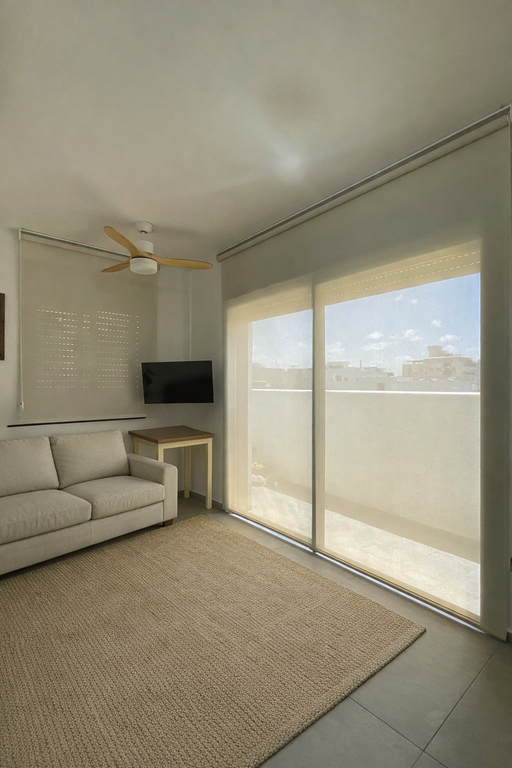 ווילון גלילה חצי שקוף לסלון עם יציאה למרפסת, מאוורר תקרה מעץ וספה בהירה.