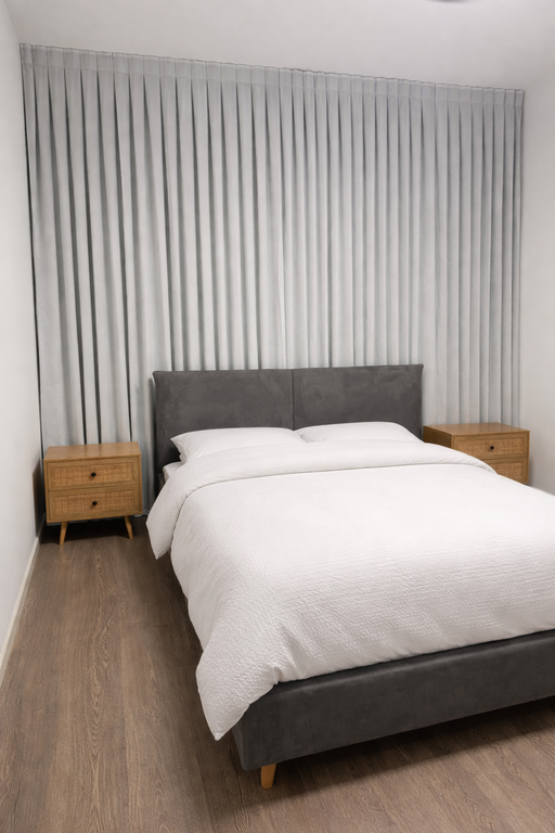 וילון בד אפור בהיר מאחורי מיטה זוגית עם גב מיטה מאפור ושידות עץ.