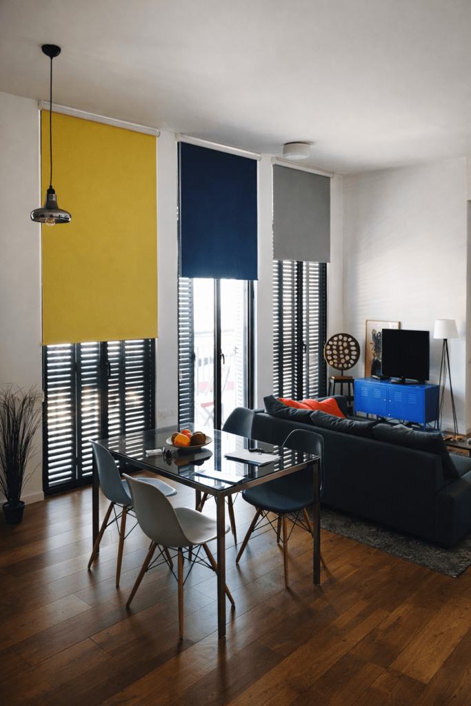 שלושה וילונות גלילה בצבעים צהוב, כחול ואפור מעל דלתות זכוכית עם תריסי שלבים שחורים בסלון מודרני.